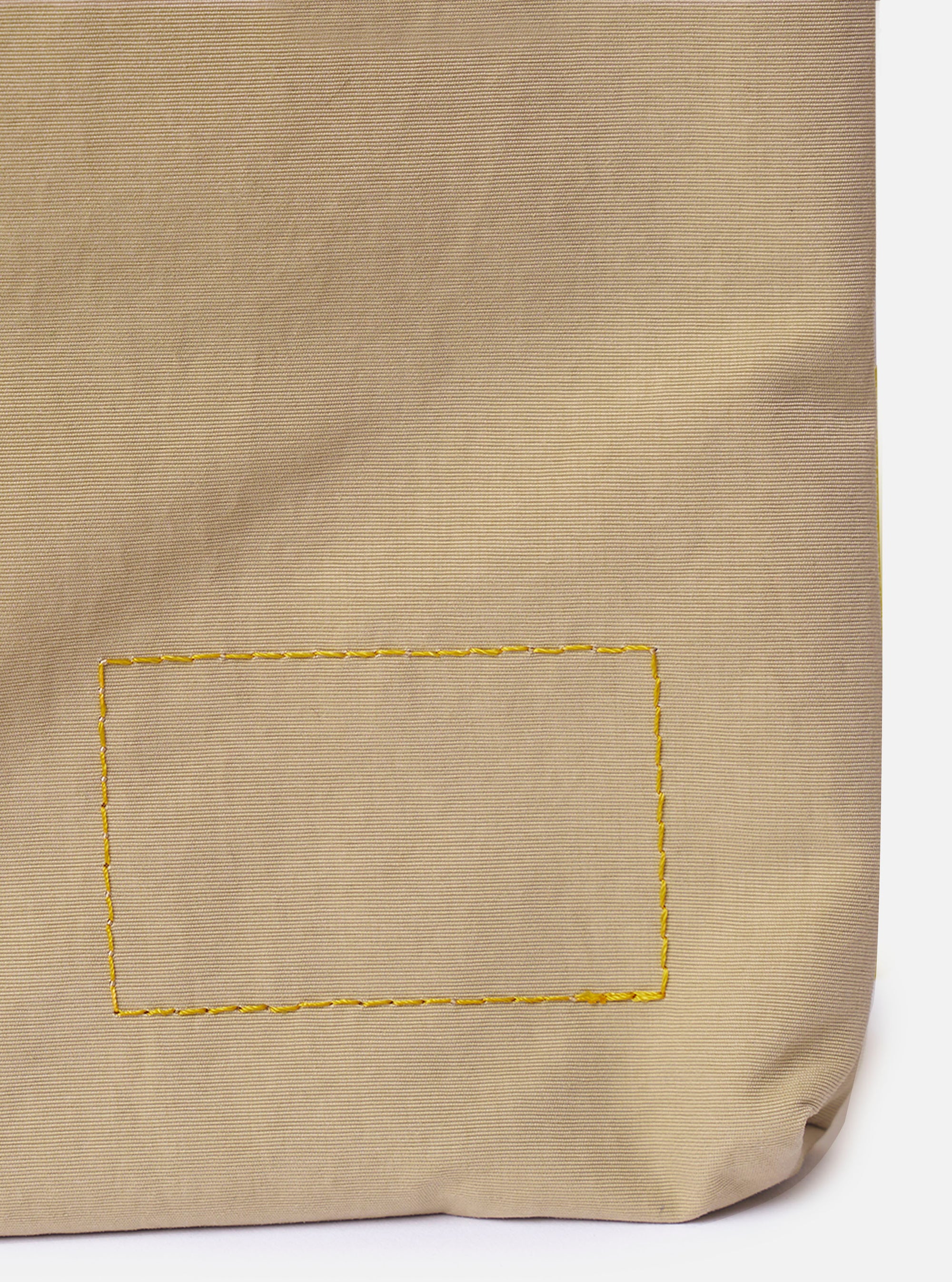 The master-piece x Universal Works Beige Canvas Sacoche is shown from a rear angle as a flat lay against a plain white background. The image highlights the adjustable olive green webbing shoulder strap and the back of the bag, which features a clean, rectangular silhouette in beige canvas.