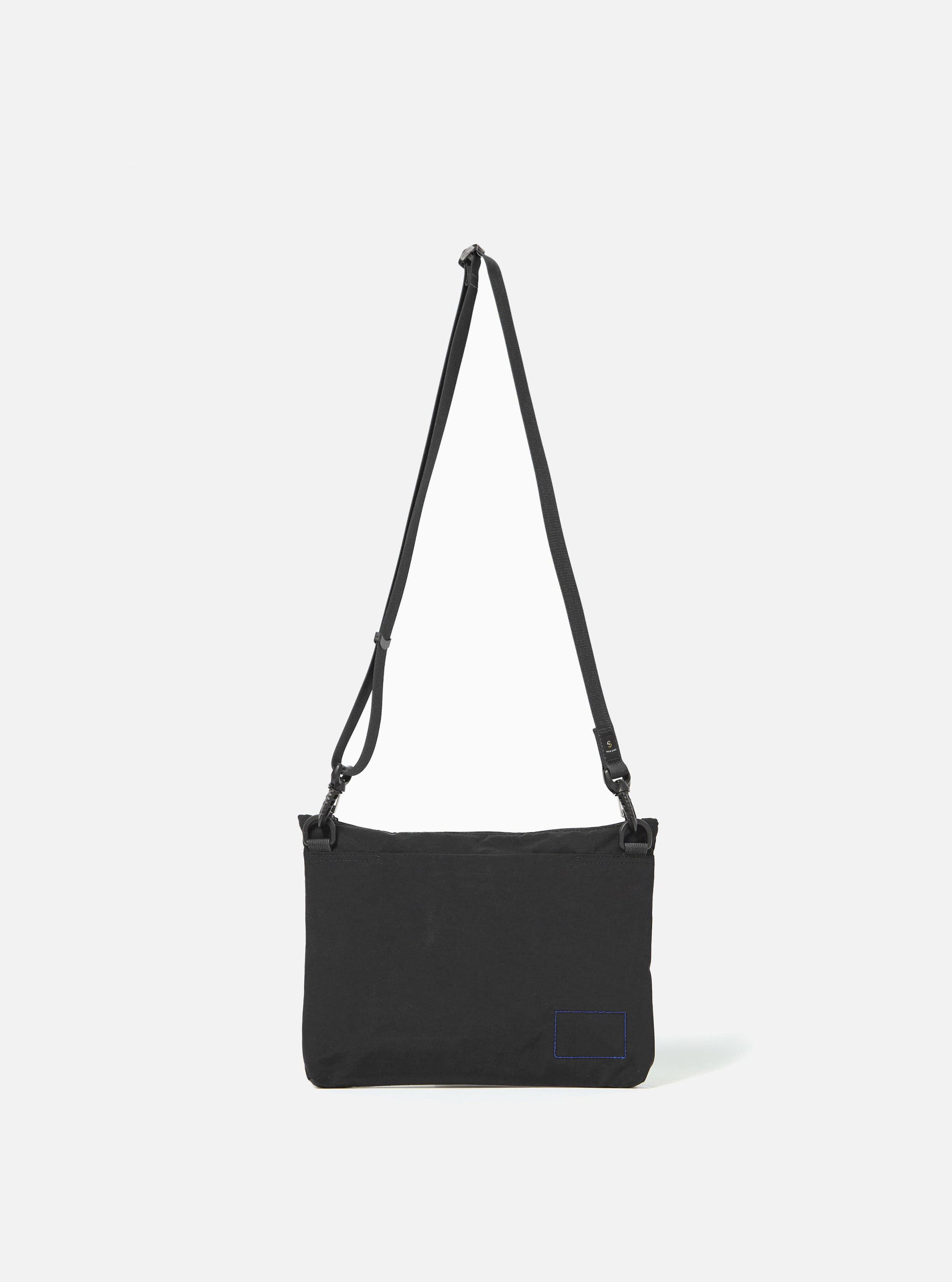 The master-piece x Universal Works Black Canvas Sacoche is shown from a rear angle as a flat lay against a white background. The image highlights the adjustable black shoulder strap attached with metal clips, a small brand tag near the bottom seam, and the clean, rectangular construction of the bag.