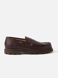 Dark brown smooth leather penny loafer shot in profile against a light grey studio backdrop. The saddle strap sits across the vamp with a central slot detail. A small green Paraboot woven label is stitched to the quarter. Visible contrast cream welt stitching runs along the chunky dark rubber lug sole. Tan leather insole lining visible at the collar.