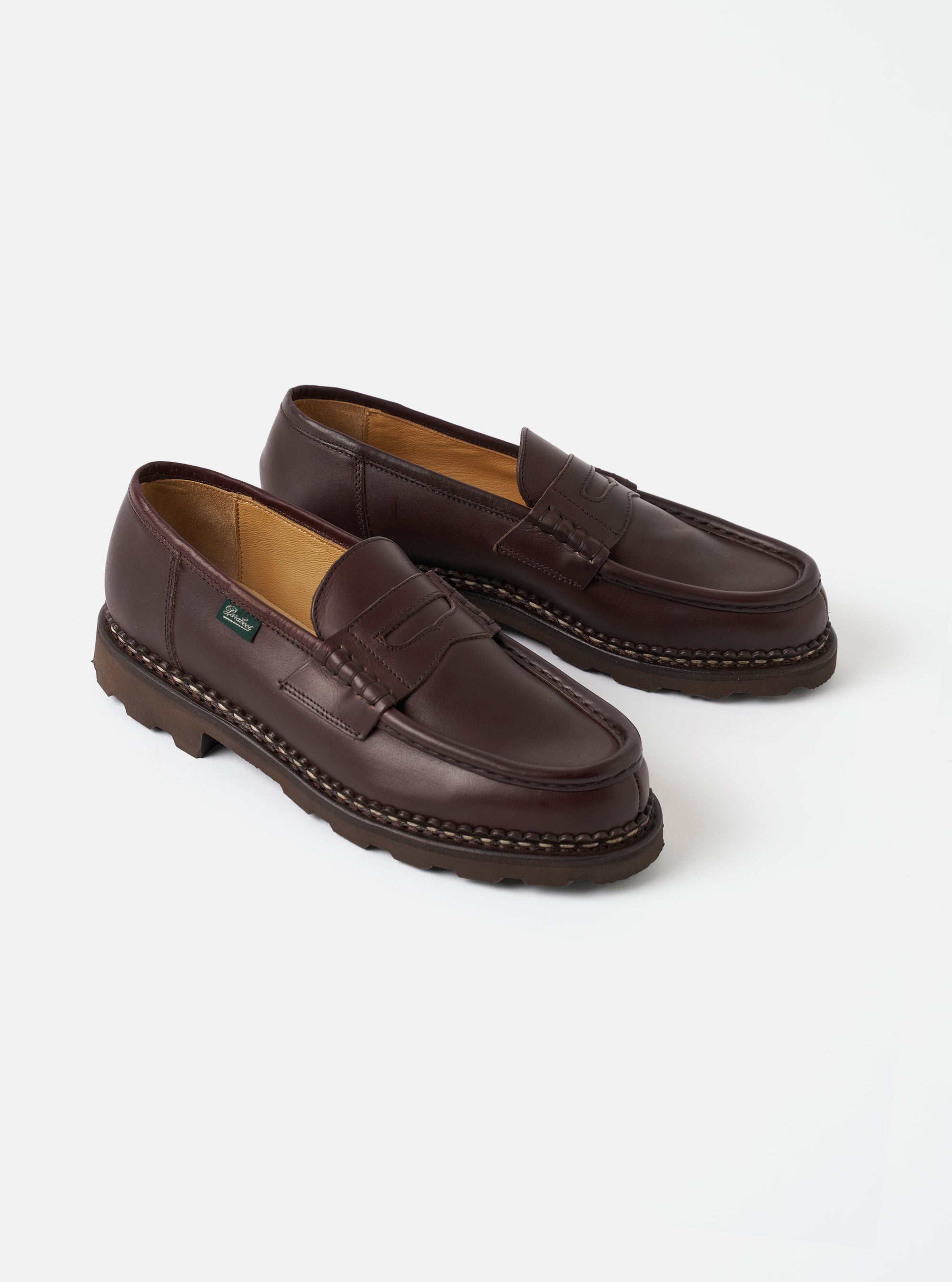 Pair of dark brown leather penny loafers on a plain light grey studio surface. Smooth cafe leather upper with a classic penny strap across the vamp and apron toe. Tan leather lining visible at the opening. Contrast braided welt stitching and a chunky dark rubber lug sole. Small green branded label on the outer side wall.