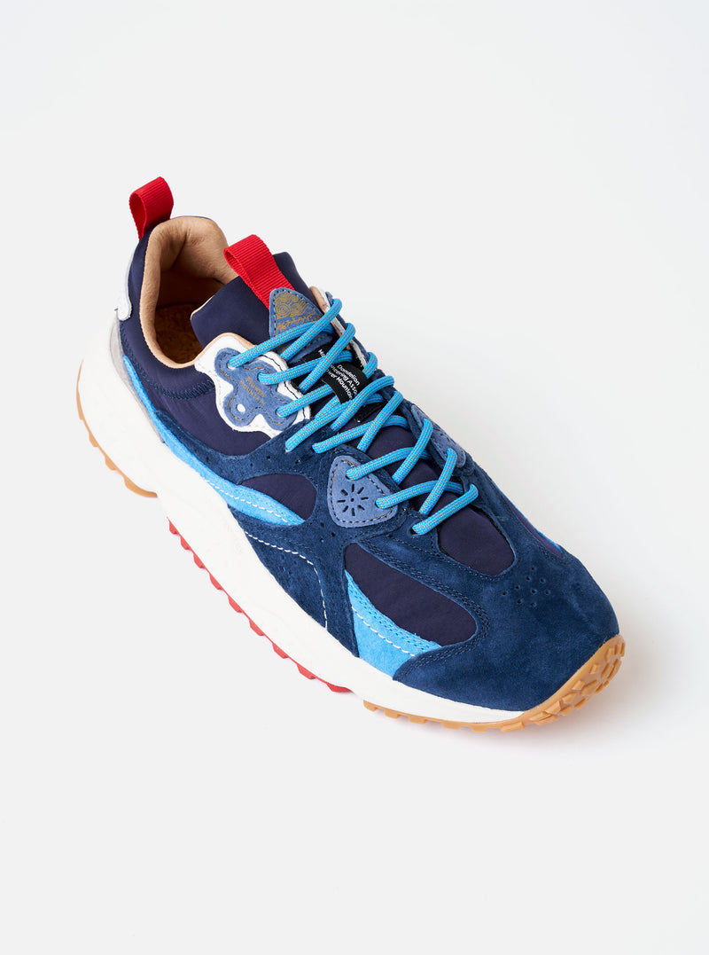 The Flower Mountain Navy/Azure Suede/Nylon Wave Man sneaker is shown from a side profile as a flat lay against a white background. The low-top shoe features a navy blue technical fabric upper with overlapping azure blue and dark navy suede overlays. It includes a classic lace-up system with trekking-style laces, a heel pull tab, and a signature notched rubber gum sole in a light tan color.