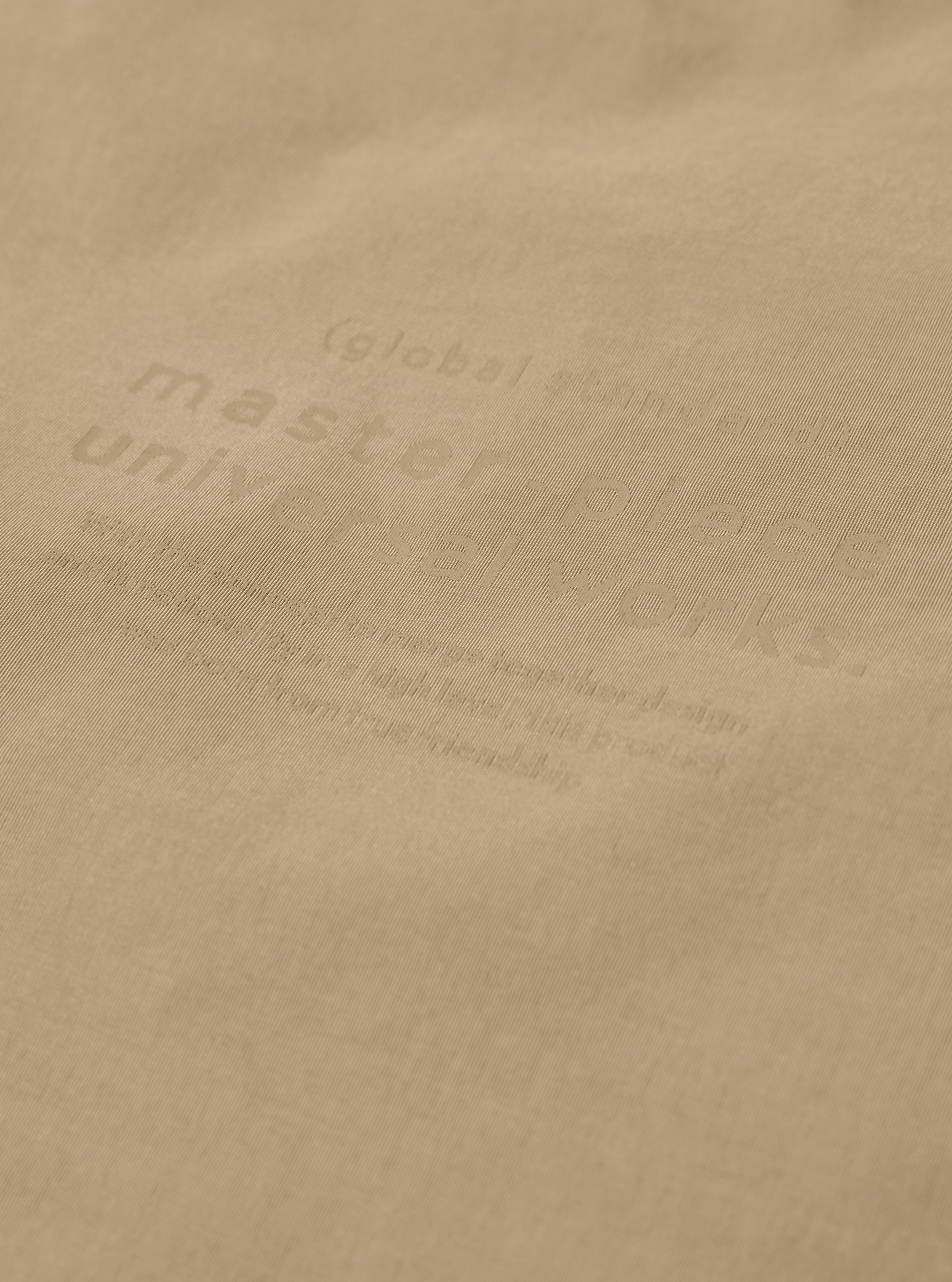 The master-piece x Universal Works Beige Large Canvas Shoulder Bag is shown from the back as a flat lay against a white background. The image highlights the bag's textured beige canvas fabric, a small black brand tag, and the adjustable black shoulder strap.