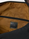 The master-piece x Universal Works Black Large Canvas Shoulder Bag is shown from a rear three-quarter angle as a flat lay against a plain white background. The image highlights the bag's large rectangular shape, the durable black canvas construction, and a side-entry zipper. A black adjustable shoulder strap is attached to the top corners, and a small collaborative brand tag is visible near the bottom edge.