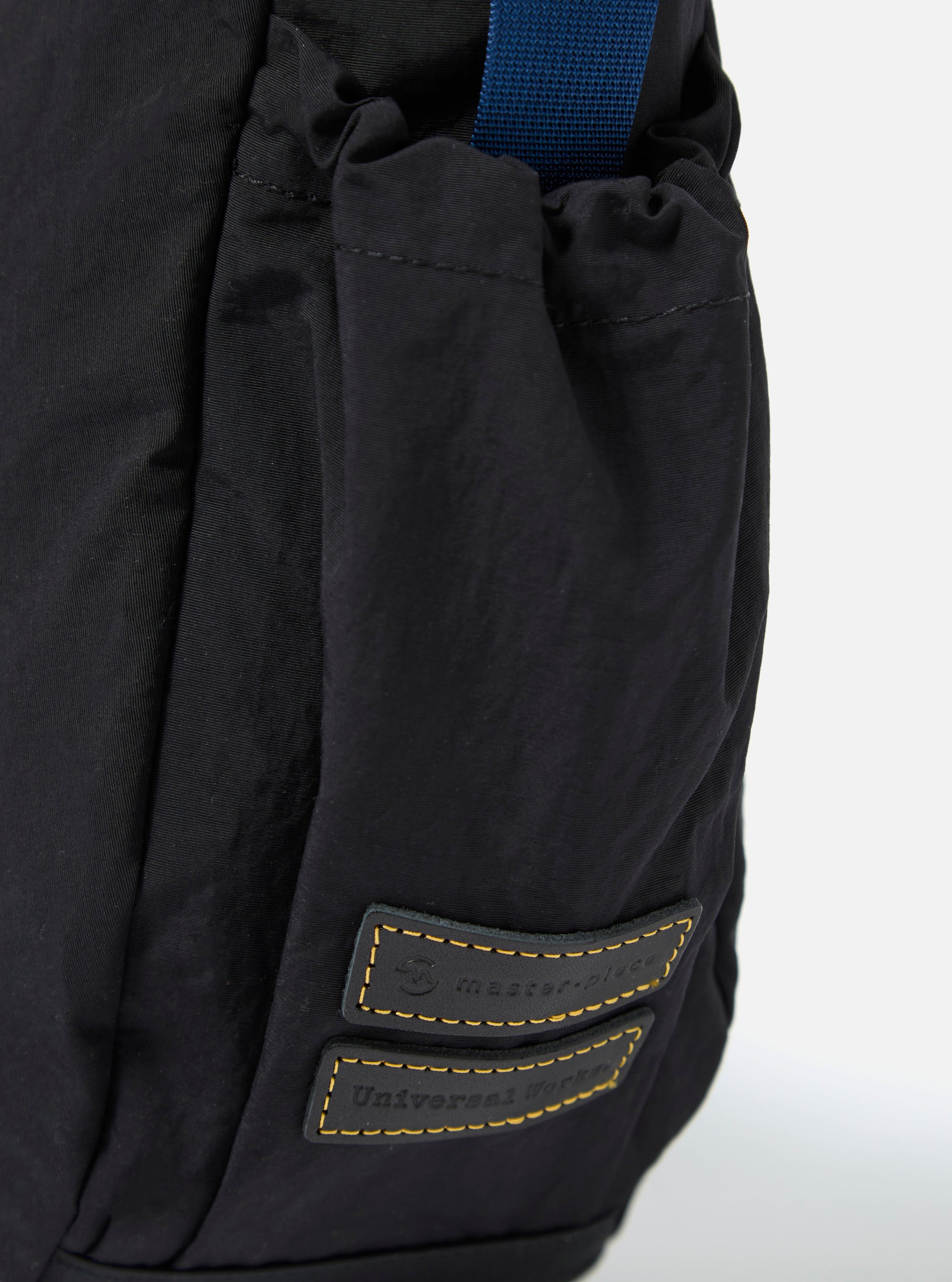 The master-piece x Universal Works Black Medium Canvas Shoulder Bag is shown from the back as a flat lay against a plain white background. The image highlights the clean, rectangular construction of the bag and the smooth texture of the black canvas fabric. A small black brand tag is visible on the side.