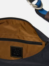 The master-piece x Universal Works Black Medium Canvas Shoulder Bag is shown from the back as a flat lay against a plain white background. The image highlights the clean, durable black canvas construction, featuring a horizontal strap across the center for attaching to a suitcase handle. A small, dual-branded logo patch is visible near the bottom edge.