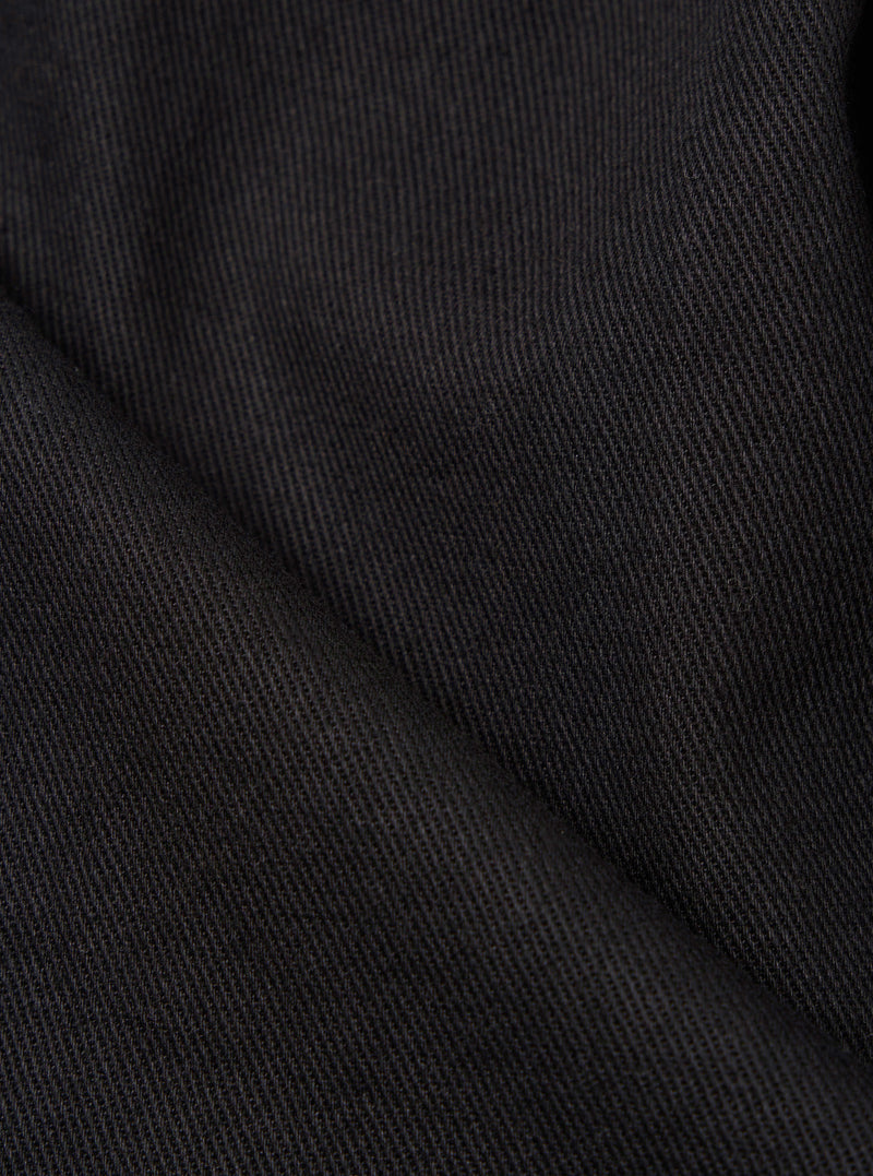 Close-up of the Black Twill Double Pleat Pant. The dark twill fabric is folded, showing the texture and weave. Shot on a plain surface.