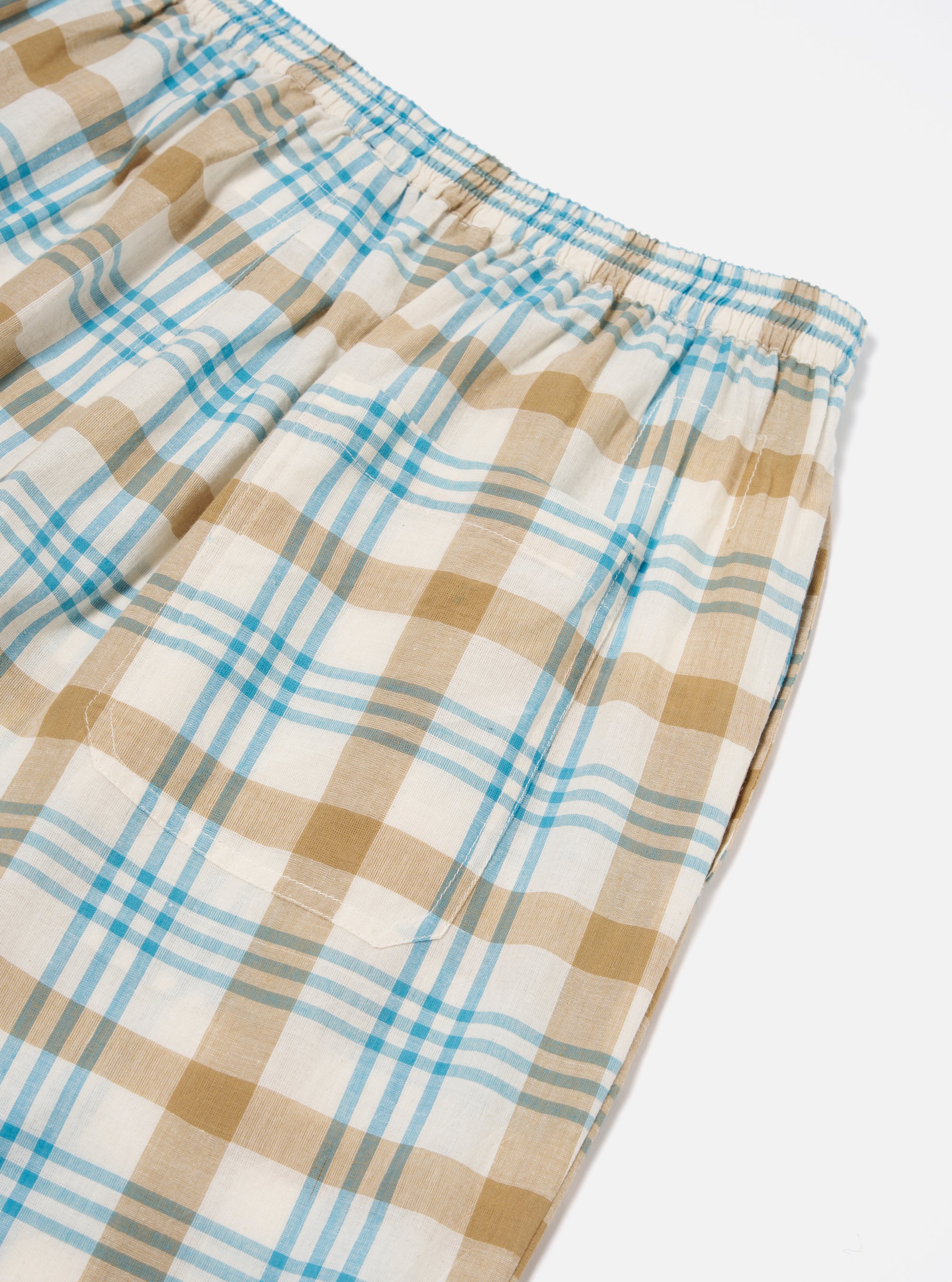 Close-up of the Ecru Madras Check Pyjama Pant on a white surface. The relaxed fit pant features a blue and tan plaid pattern and an elastic waistband.