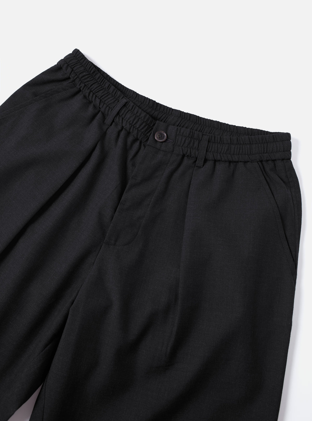 Close-up of the Charcoal Tropical Suiting Oxford Pant on a white surface. The charcoal pants have an elastic waistband and a button closure.