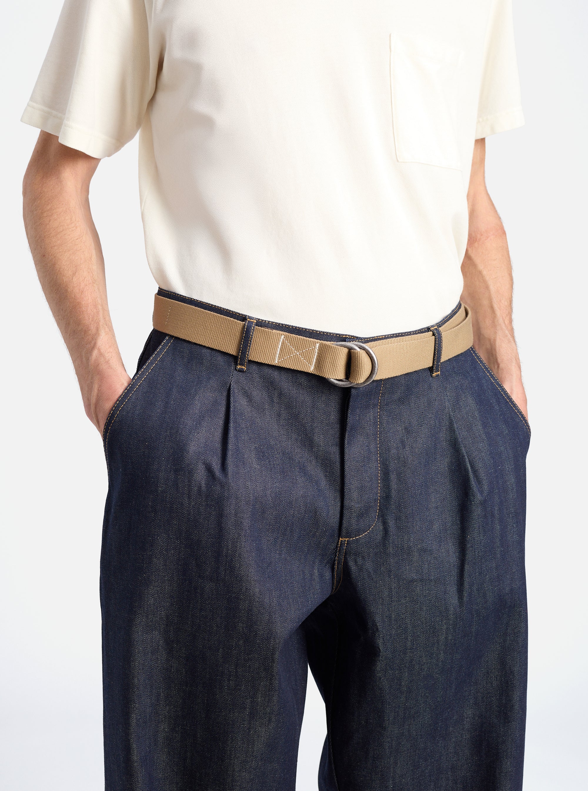 A model wears a Dark Sand Canvas O Ring Belt with dark denim pants against a white background. The belt is canvas with a silver O-ring buckle.