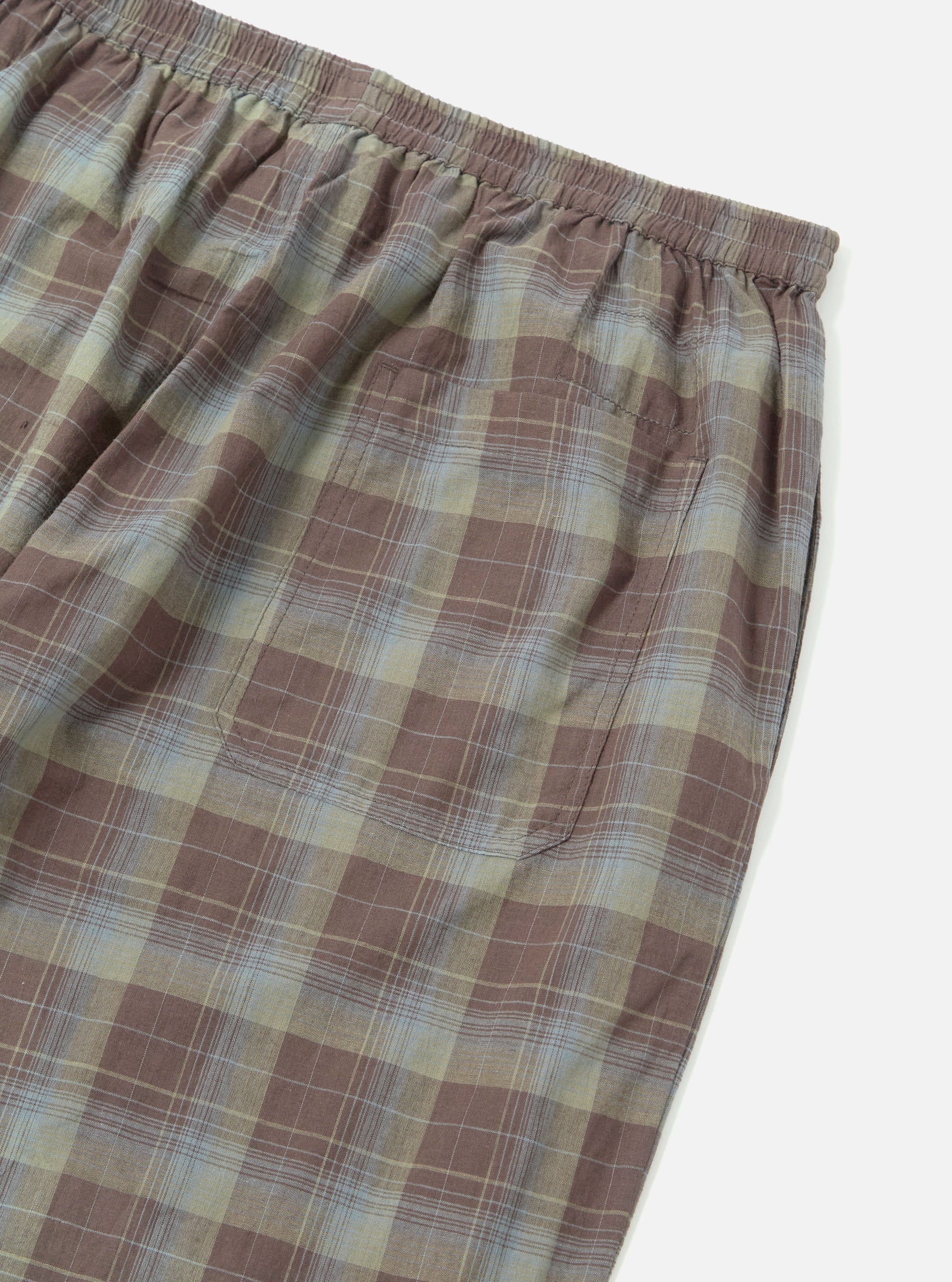 Close-up of the Green/Brown Check Pyjama Pant on a white surface. The fabric shows a plaid pattern with brown, green, and light blue lines. Elastic waistband and pocket detail visible.