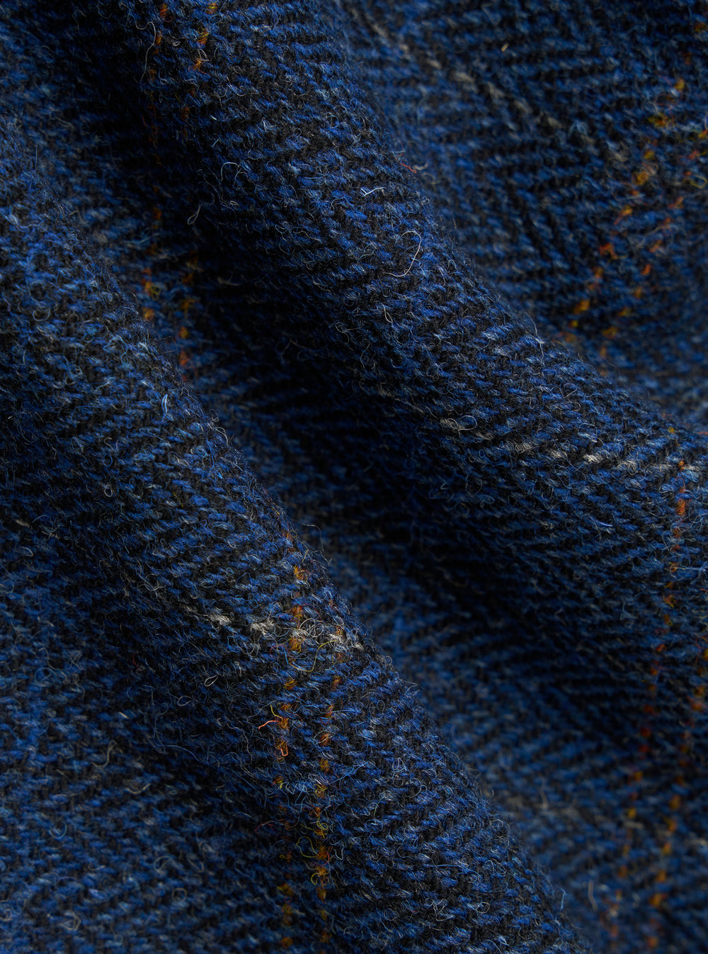 Close-up of the Navy Harris Tweed Plaid Bakers Jacket fabric. The blue and black herringbone tweed has subtle orange accents. The material is softly draped.