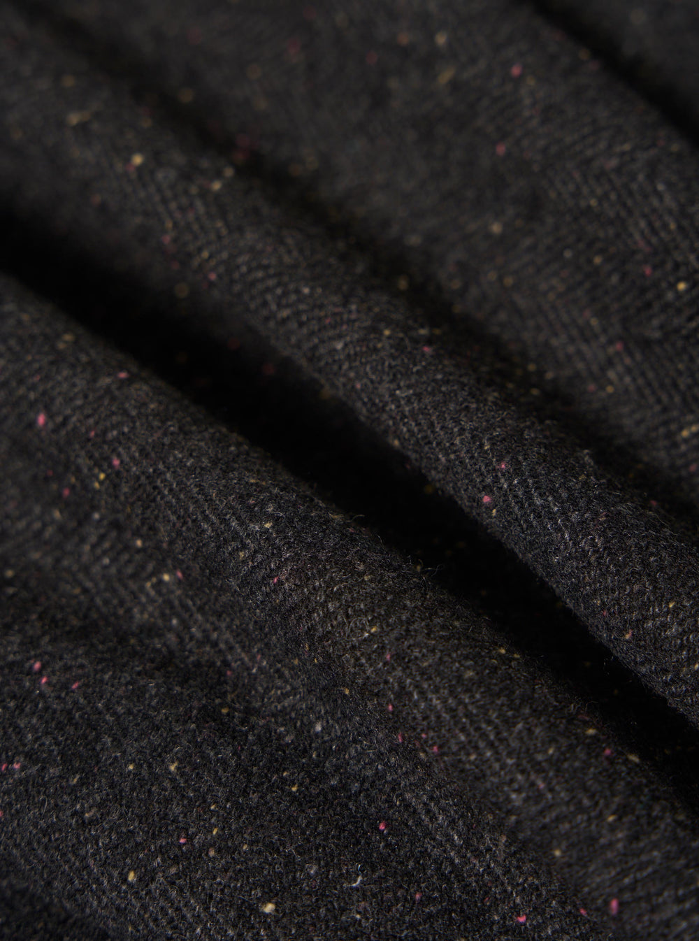 Close-up of the Black Salt N Pepper Tweed Station Jacket fabric. The tweed is a dark color with flecks of pink and yellow. The fabric is draped to show texture.