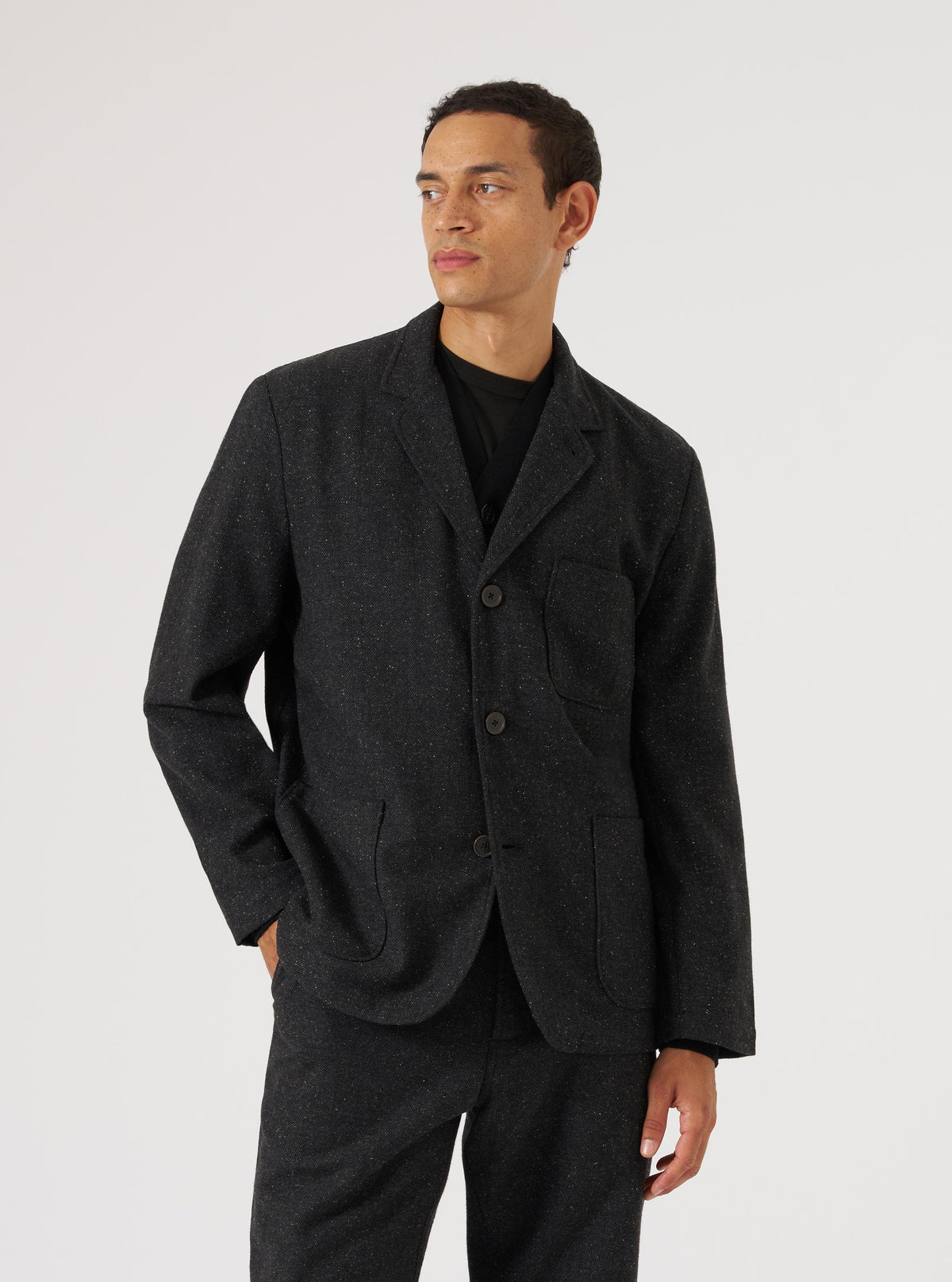 A model poses in a Black Salt N Pepper Tweed Three Button Jacket against a white backdrop. The tweed jacket has a classic fit and visible buttons.