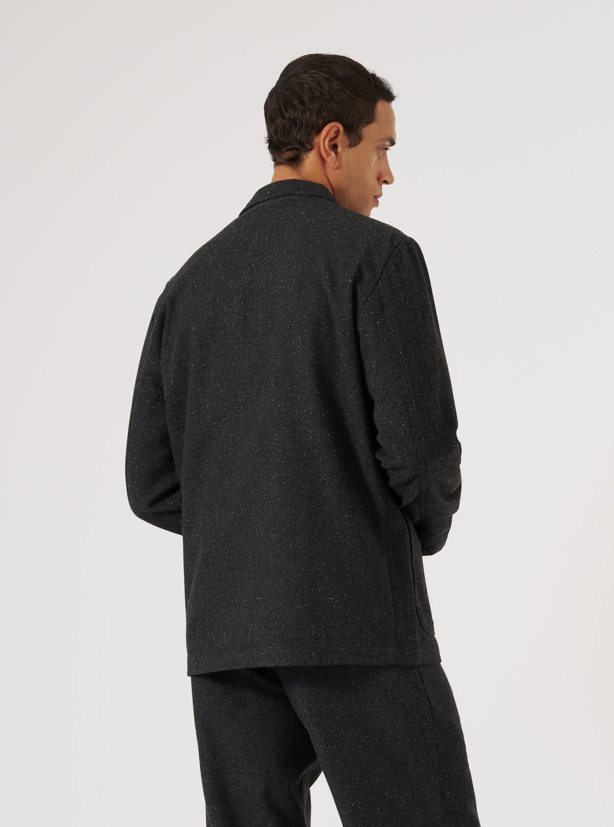 A man models the Black Salt N Pepper Tweed Three Button Jacket against a white backdrop. The jacket has a tailored fit and visible tweed texture.