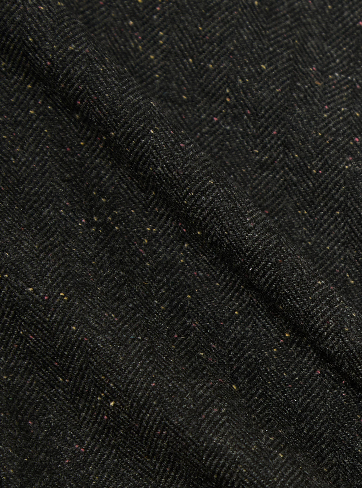 Close-up of the Black Salt N Pepper Tweed Three Button Jacket. The tweed fabric has a subtle herringbone pattern with flecks of yellow and pink. The background is a plain surface.