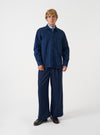 A male model stands against a plain neutral backdrop wearing the Indigo Work Denim Fatigue Shirt. He wears it unbuttoned over a plain t-shirt, highlighting the classic fatigue utility pockets, crisp collar, and rich indigo denim fabric.