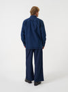 A male model seen from the side wearing the Indigo Work Denim Fatigue Shirt against a plain light studio background. The shot highlights the relaxed fit, long sleeves, sturdy cotton denim texture, button cuffs, and neat straight hem.