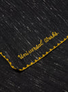 Close-up of a Black Wool Scarf with subtle white specks and gold embroidered trim and text on a flat surface.