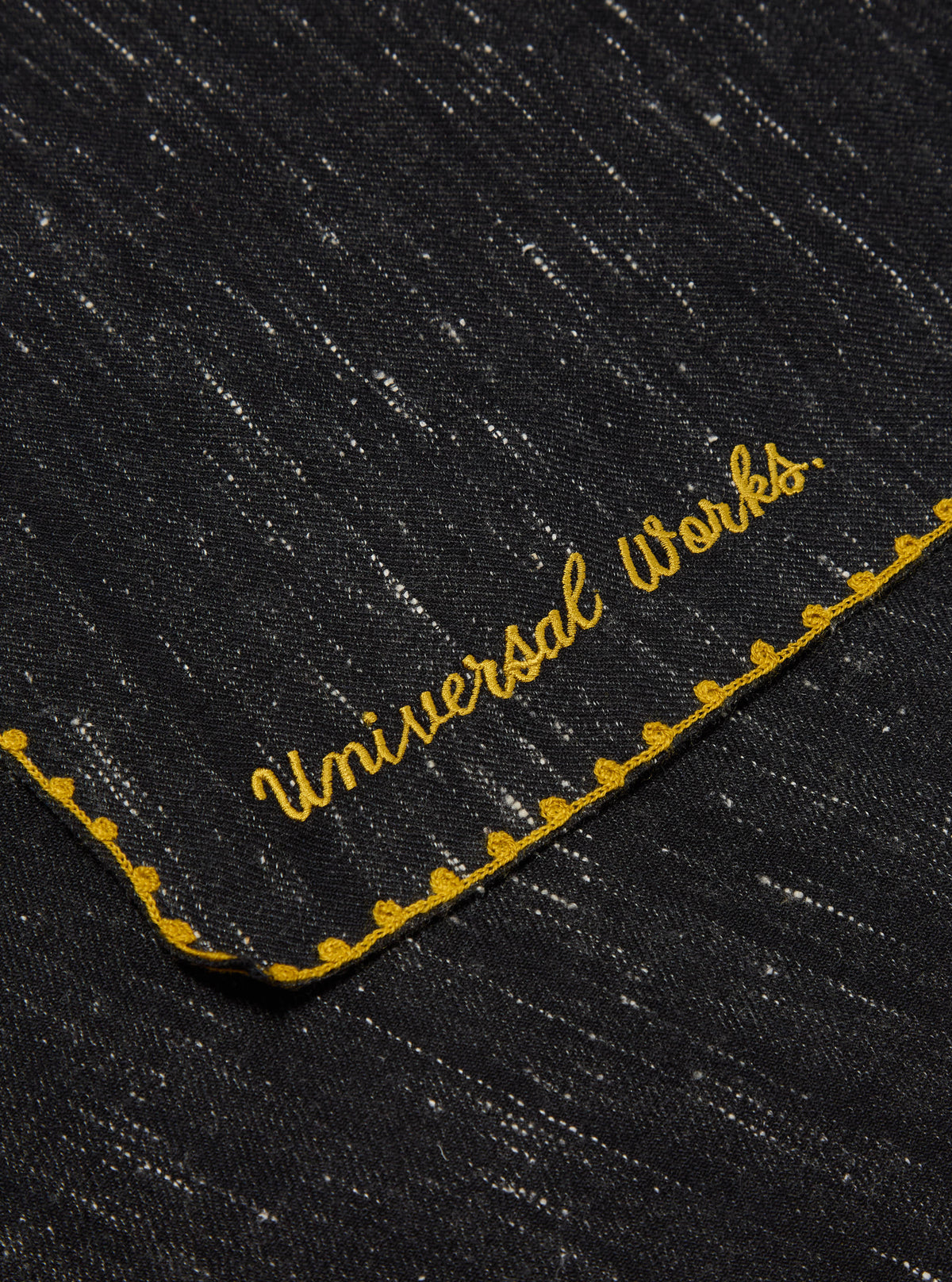 Close-up of a Black Wool Scarf with subtle white specks and gold embroidered trim and text on a flat surface.