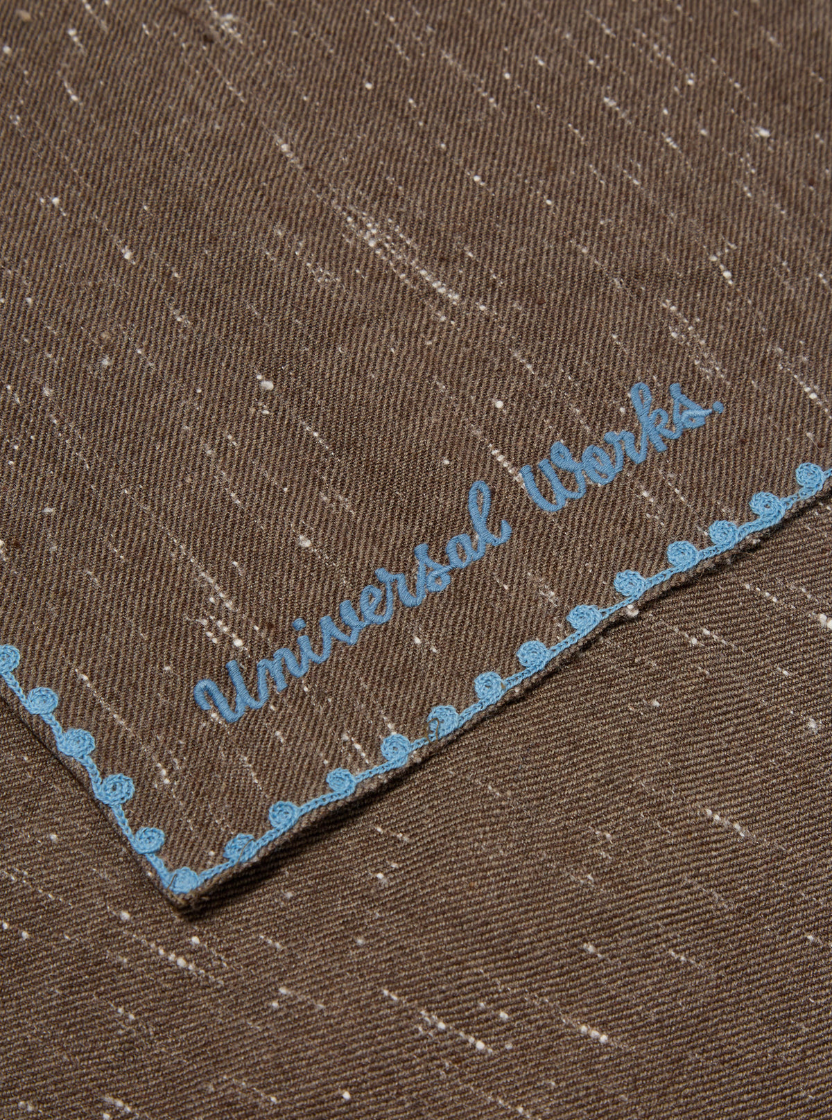 Close-up of a Brown Wool Scarf with blue embroidered trim and logo on a flat surface. The scarf has a subtle speckled pattern.