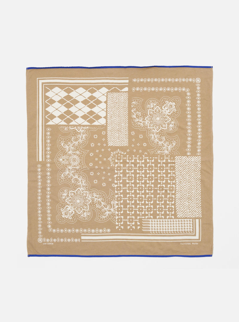 The Sand Single Jersey Neckerchief, a tan cotton square with white geometric and floral prints and a blue border, is laid flat on a white surface.