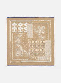 The Sand Single Jersey Neckerchief, a tan cotton square with white geometric and floral prints and a blue border, is laid flat on a white surface.