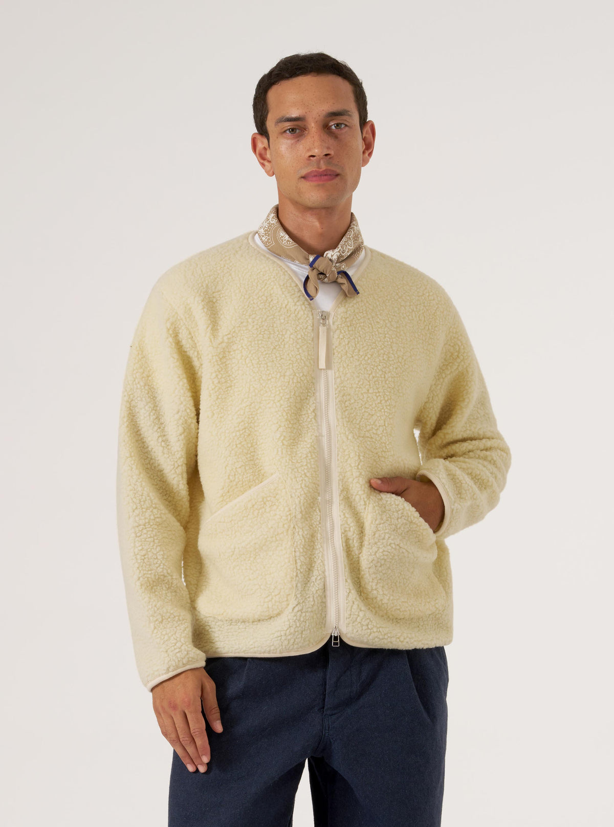 Studio shot of a model wearing a cream fleece jacket and a Sand Single Jersey Neckerchief. The neckerchief is tied loosely around the neck. White backdrop.