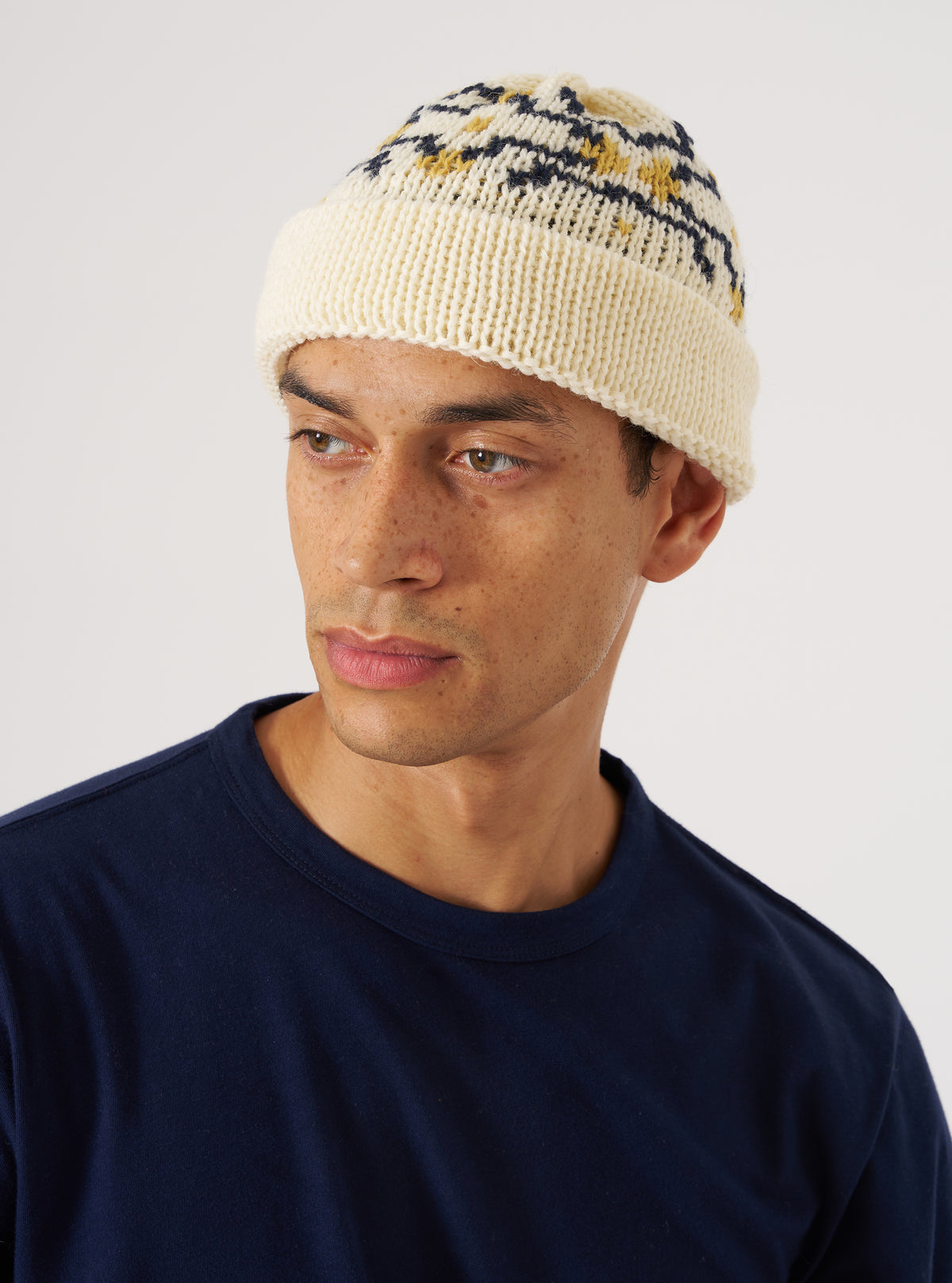 A model wears the Ecru/Navy British Wool Short Watch Cap against a white backdrop. The cap features a patterned design in ecru, navy, and gold.
