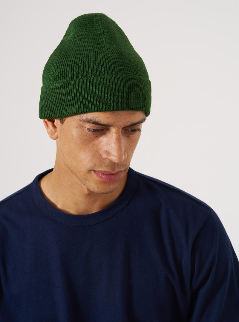 A model wears a Green Merino Knit Watch Cap against a white background. The cap is a ribbed knit, and the model is wearing a navy t-shirt.