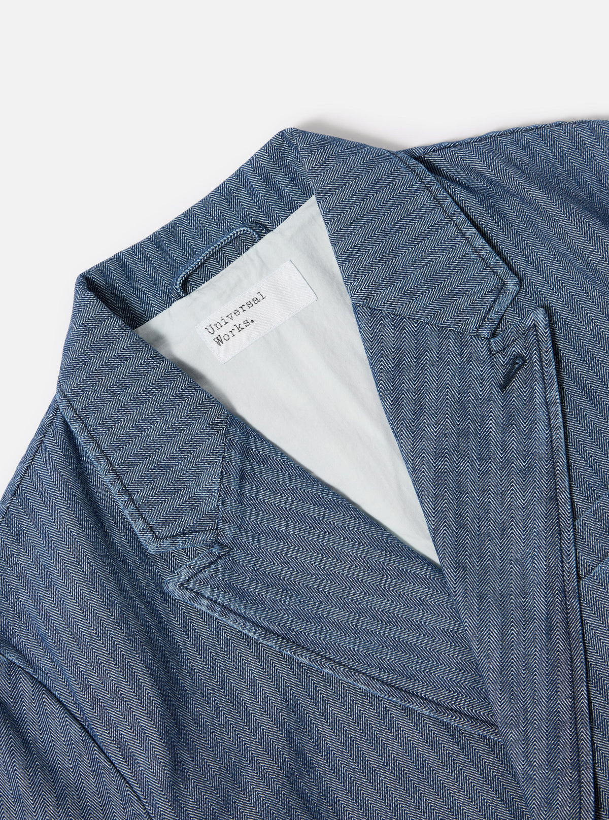 Close-up of the Indigo Denim Herringbone Manor Jacket on a white surface. The jacket features a blue and white herringbone pattern and a light-colored lining.