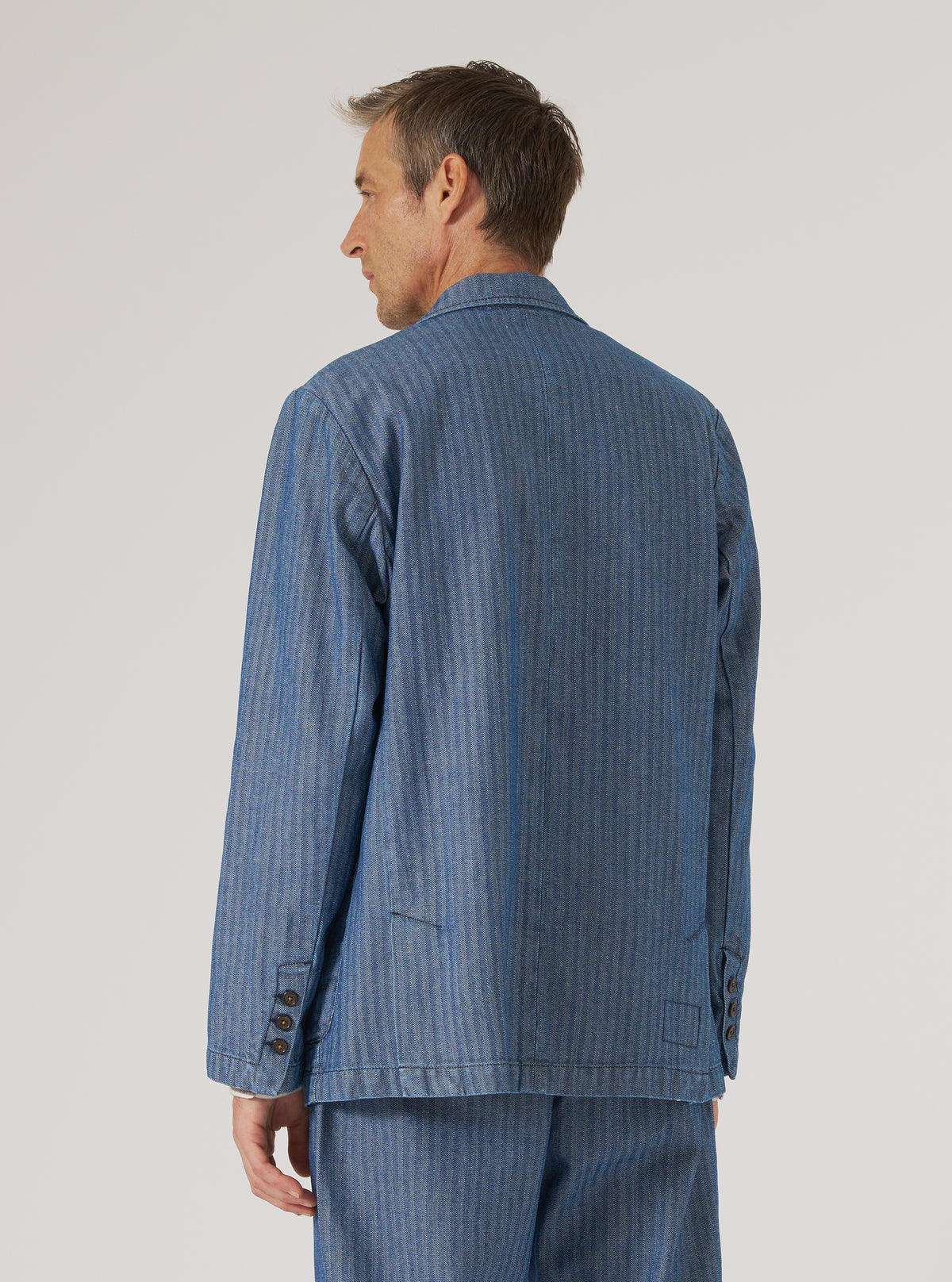 Back view of a model wearing an Indigo Denim Herringbone Manor Jacket. The blue herringbone jacket is on a man in front of a plain white background.