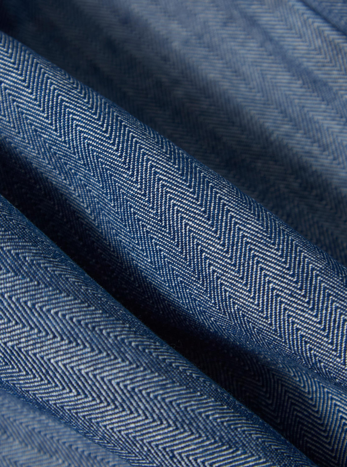 Close-up of the Indigo Denim Herringbone Manor Jacket, showcasing the blue herringbone pattern and denim texture. The fabric is draped to highlight its quality.