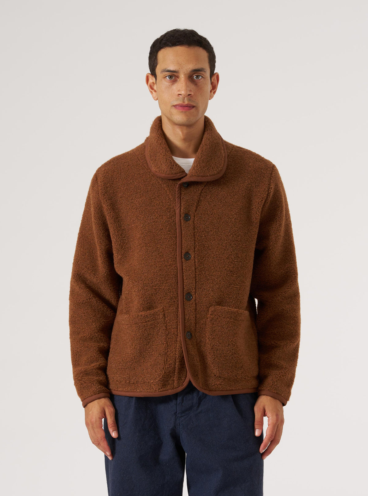 A man stands against a white backdrop, modeling the Cumin Alvar Fleece Lancaster Jacket. The brown fleece jacket features a button-up design and trim.