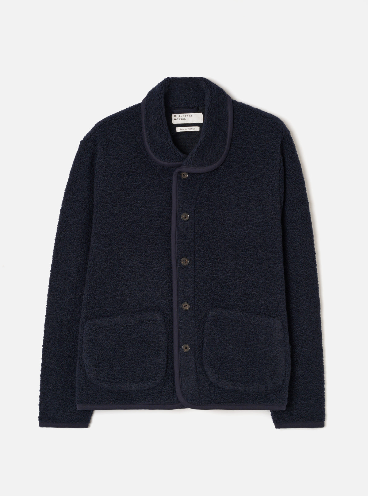 A male model stands against a plain neutral studio background wearing the Navy Alvar Fleece Lancaster Jacket. The front-view image displays the deep navy blue shade, cozy shawl collar, front button closure, and two patch pockets.