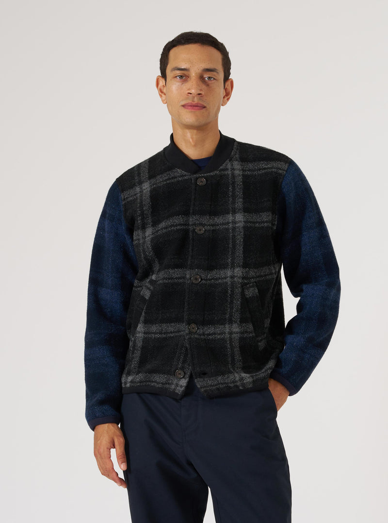 A man models the Grey Moh Check Fleece Button Front Bomber. The bomber has a grey and black check pattern on the front and blue sleeves. The background is a plain white.