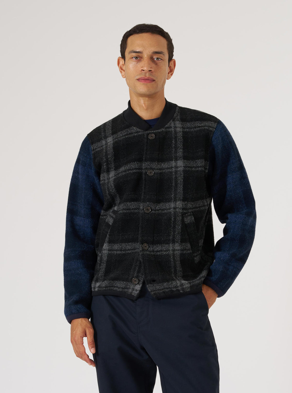 A man models the Grey Moh Check Fleece Button Front Bomber. The bomber has a grey and black check pattern on the front and blue sleeves. The background is a plain white.