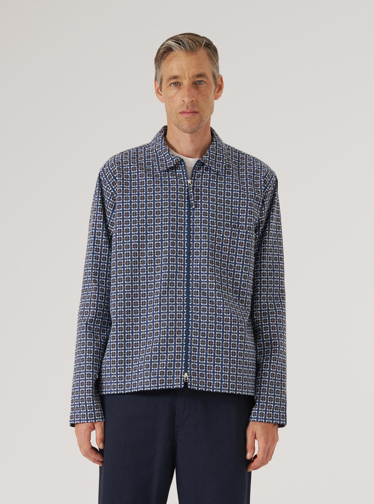 A man stands in front of a white background, wearing a Navy Oscar Cotton Zip Shirt with a geometric pattern and navy pants. The shirt is fully zipped.