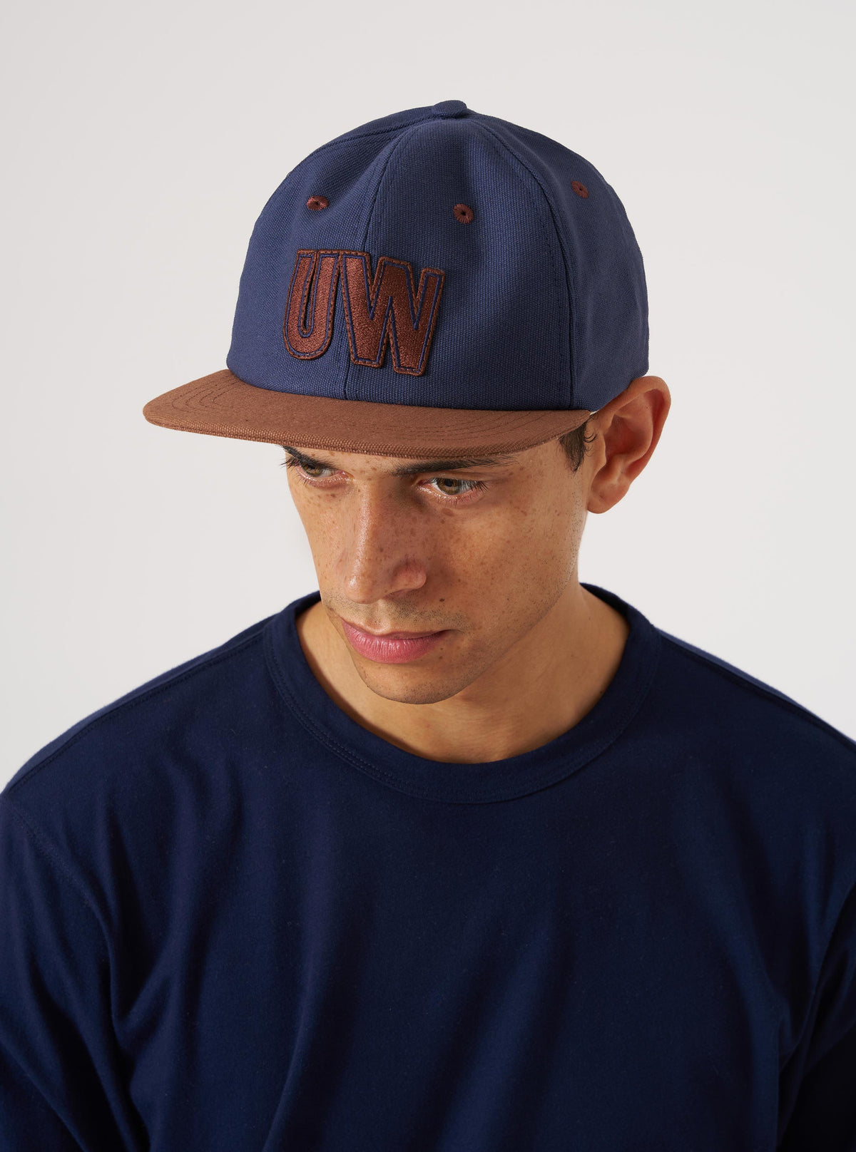 A man wears a Navy/Brown Canvas Baseball Hat in a studio shot. The hat has brown accents and a 'UW' logo. The background is a simple white backdrop.