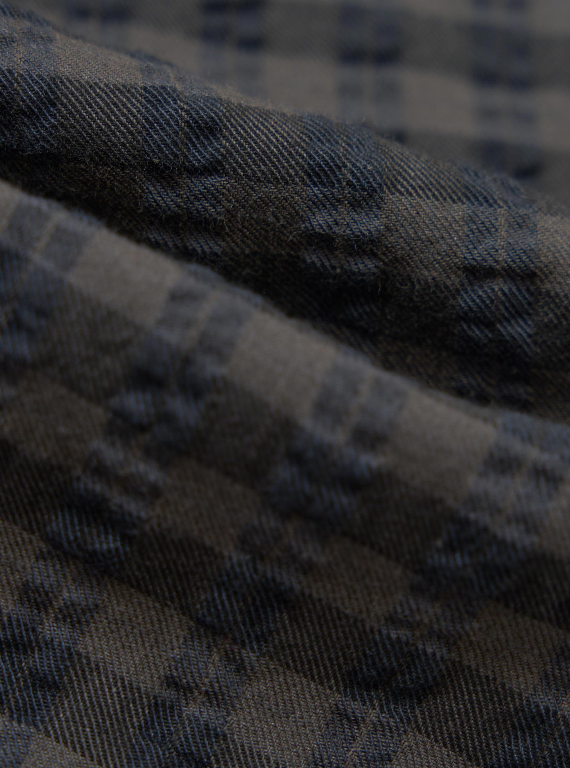 Close-up of the Charcoal/Navy Seersucker Plaid Pyjama Pant fabric. The material shows a textured plaid pattern in charcoal and navy. The fabric is folded, highlighting its soft texture.