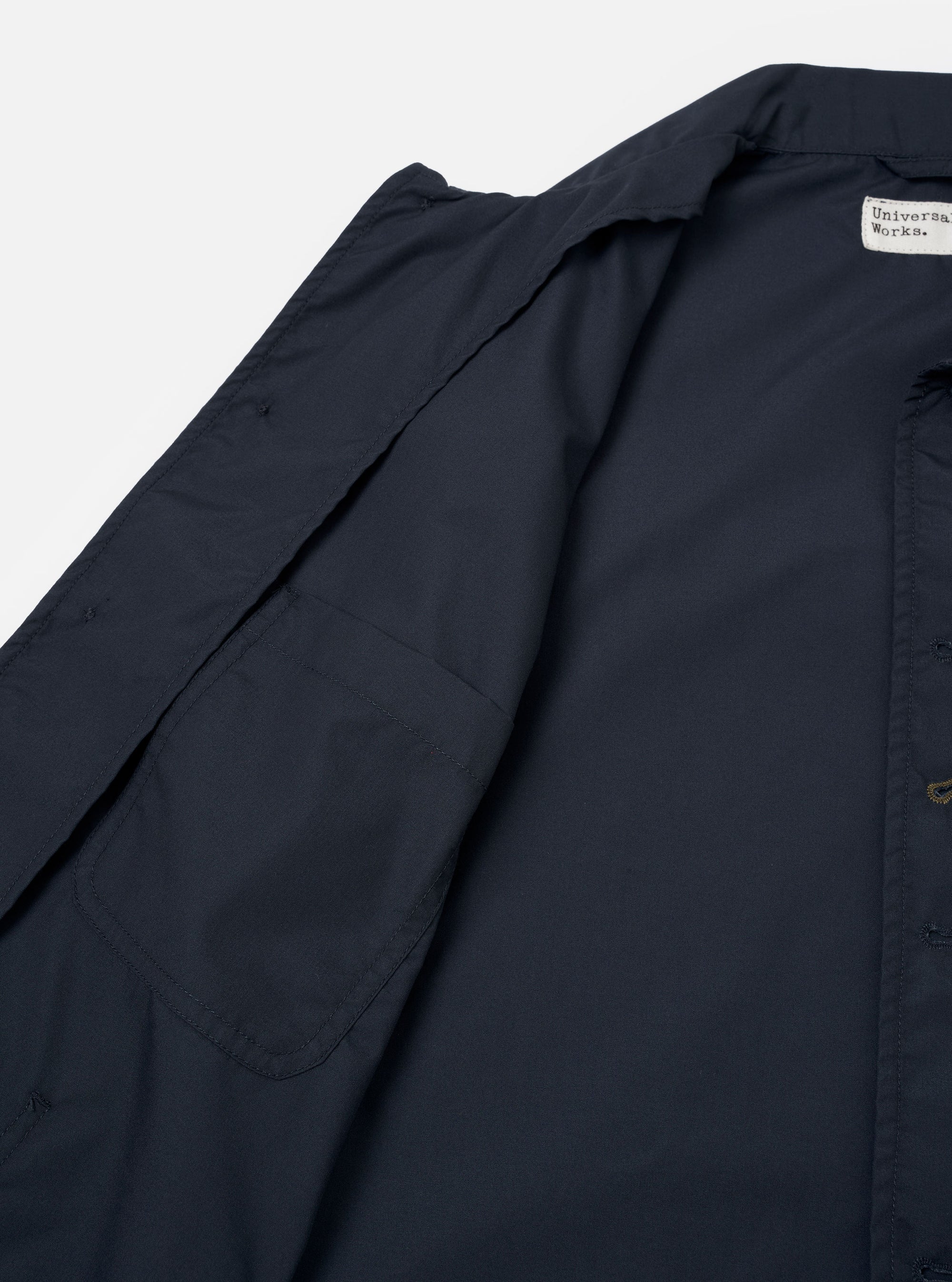 The Navy Recycled Polytech Bakers Jacket is shown in a close-up detail shot as a flat lay against a plain white background. The image focuses on the lower front corner of the jacket, showcasing a large rectangular patch pocket and the clean stitching along the hem and side. The deep navy blue fabric has a smooth, matte finish.