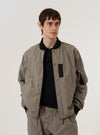 A model wears the Grey Brown Wales Glen Check NS Bomber Jacket over a dark top, standing against a plain white background. The jacket features a classic bomber silhouette with a central zip closure, a ribbed collar, and two slanted side pockets. The garment is characterized by its detailed grey and brown glen check plaid pattern throughout.