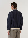 A model wears the Navy Peached Poplin NS Bomber Jacket over a light-colored top, shown from a rear three-quarter angle against a plain white background. The image highlights the jacket's articulated sleeves, contrast stripe ribbing on the collar and cuffs, and the smooth texture of the navy peached poplin fabric. The jacket is shown with a regular fit and a straight hem.