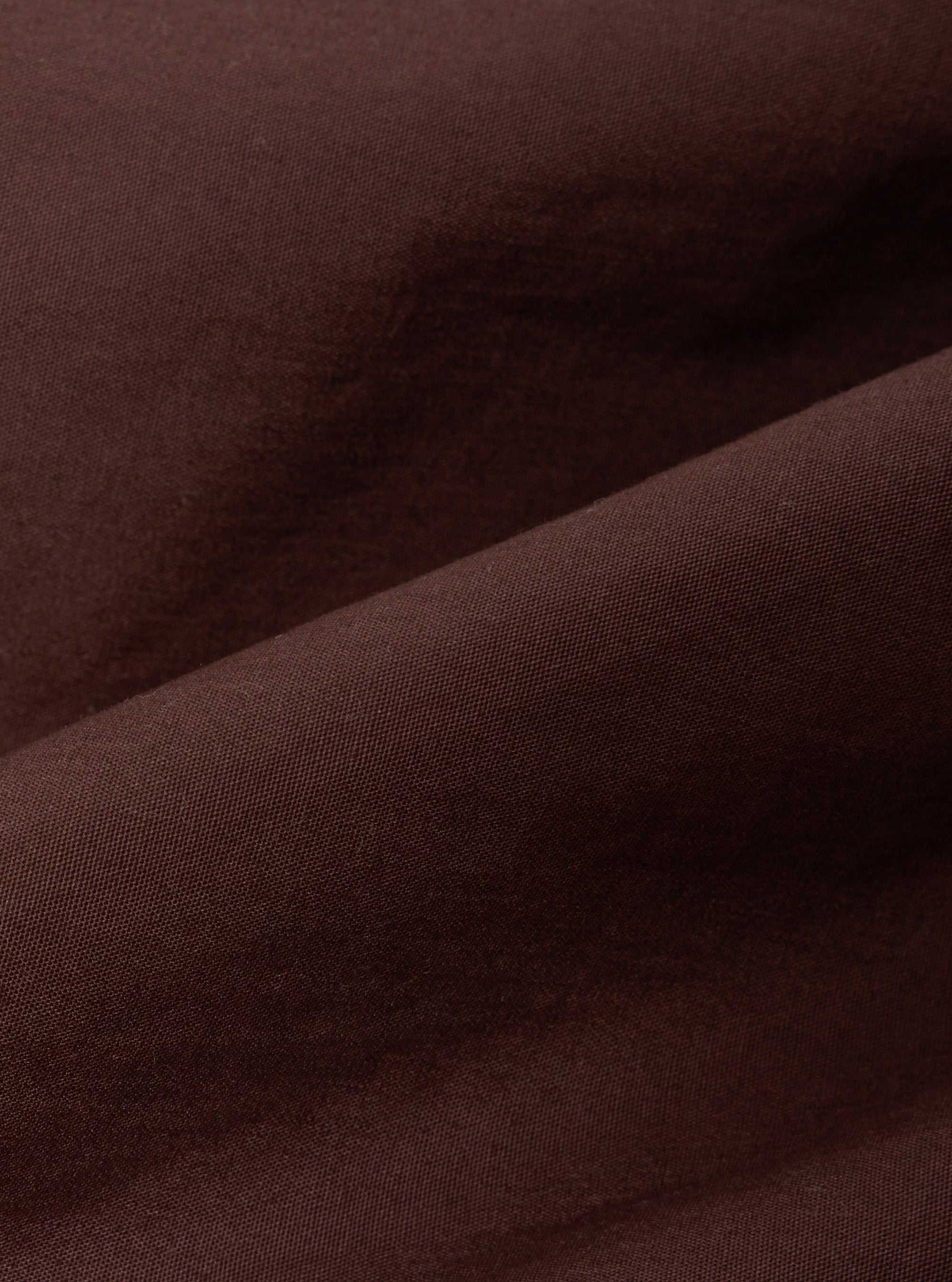 A close-up fabric swatch of the Brown Peached Poplin NS Bomber Jacket highlights the smooth, matte texture and rich brown color of the peached poplin material. The fabric fills the frame, providing a detailed view of the garment's weave and solid color.