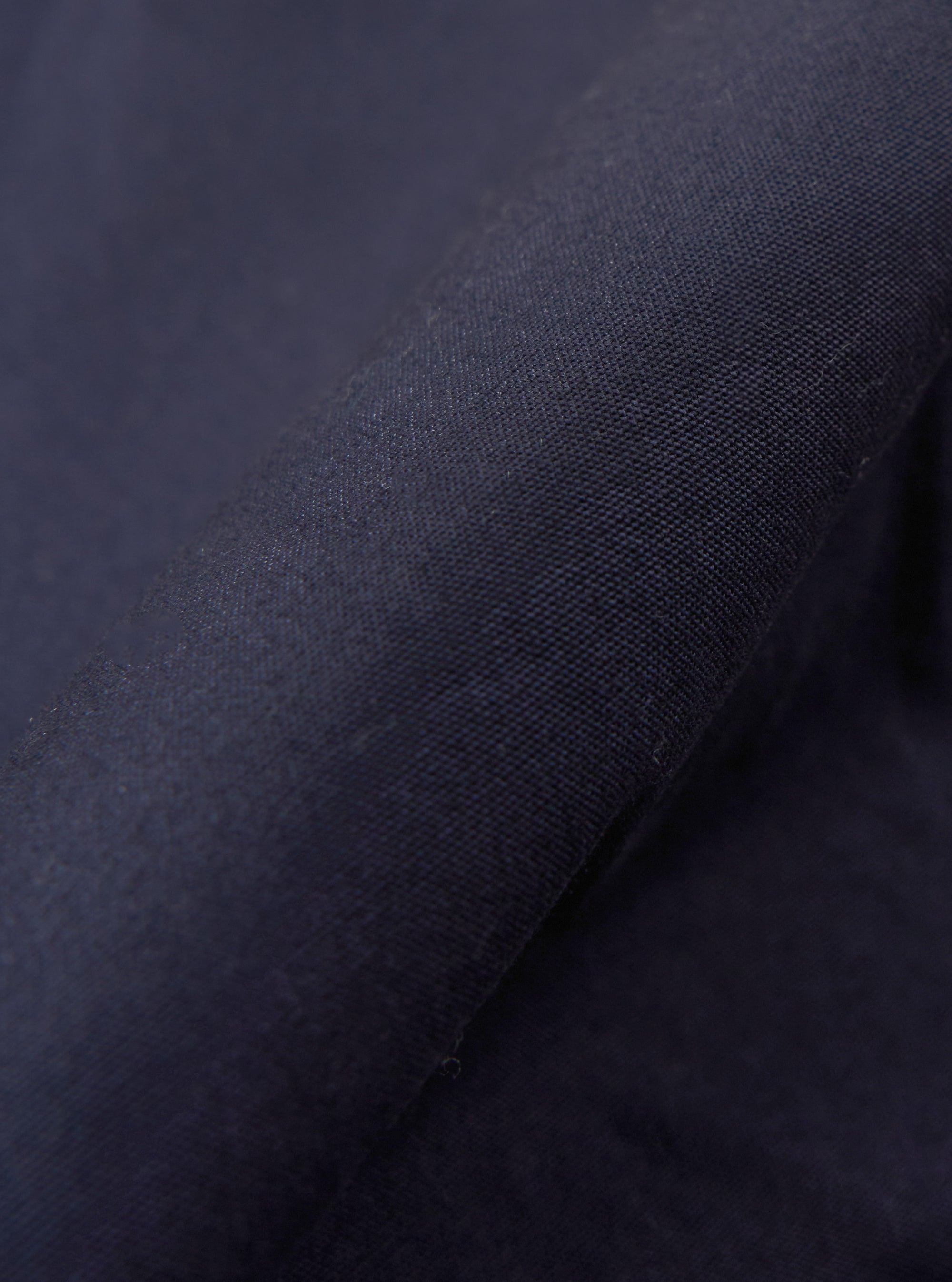 A close-up fabric swatch of the Navy Peached Poplin NS Bomber Jacket highlights the smooth, matte texture and deep navy blue color of the peached poplin material. The fabric fills the frame, providing a detailed view of the garment's weave and finish.