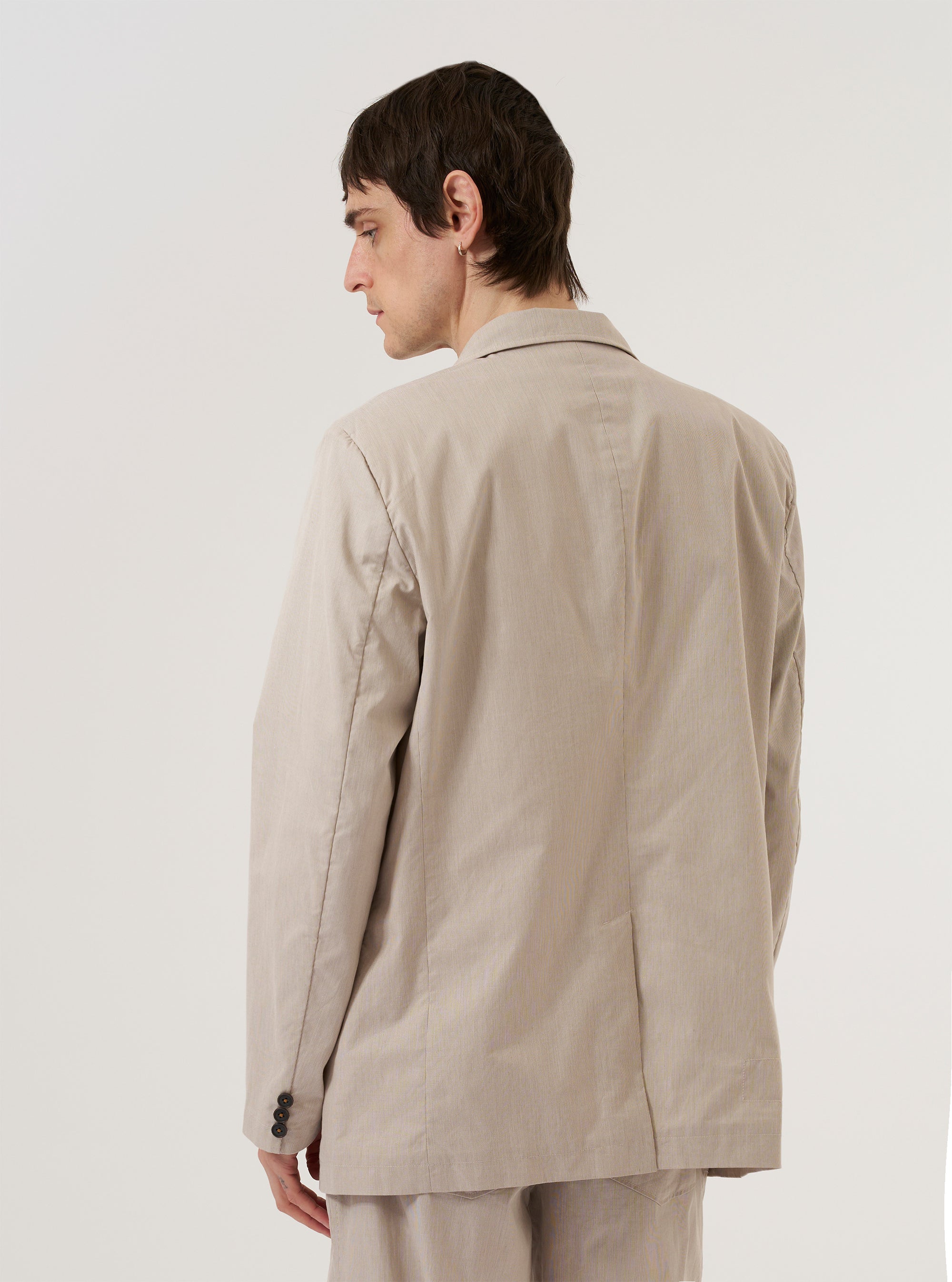 A model wears the Brown Kennedy Stripe Boston Blazer over a neutral-colored shirt, shown from a rear three-quarter angle against a plain, light grey background. The blazer features a vertical striped pattern in shades of brown and white, a structured back with a single central vent, and a relaxed tailored fit. The texture of the cotton fabric is visible under studio lighting.