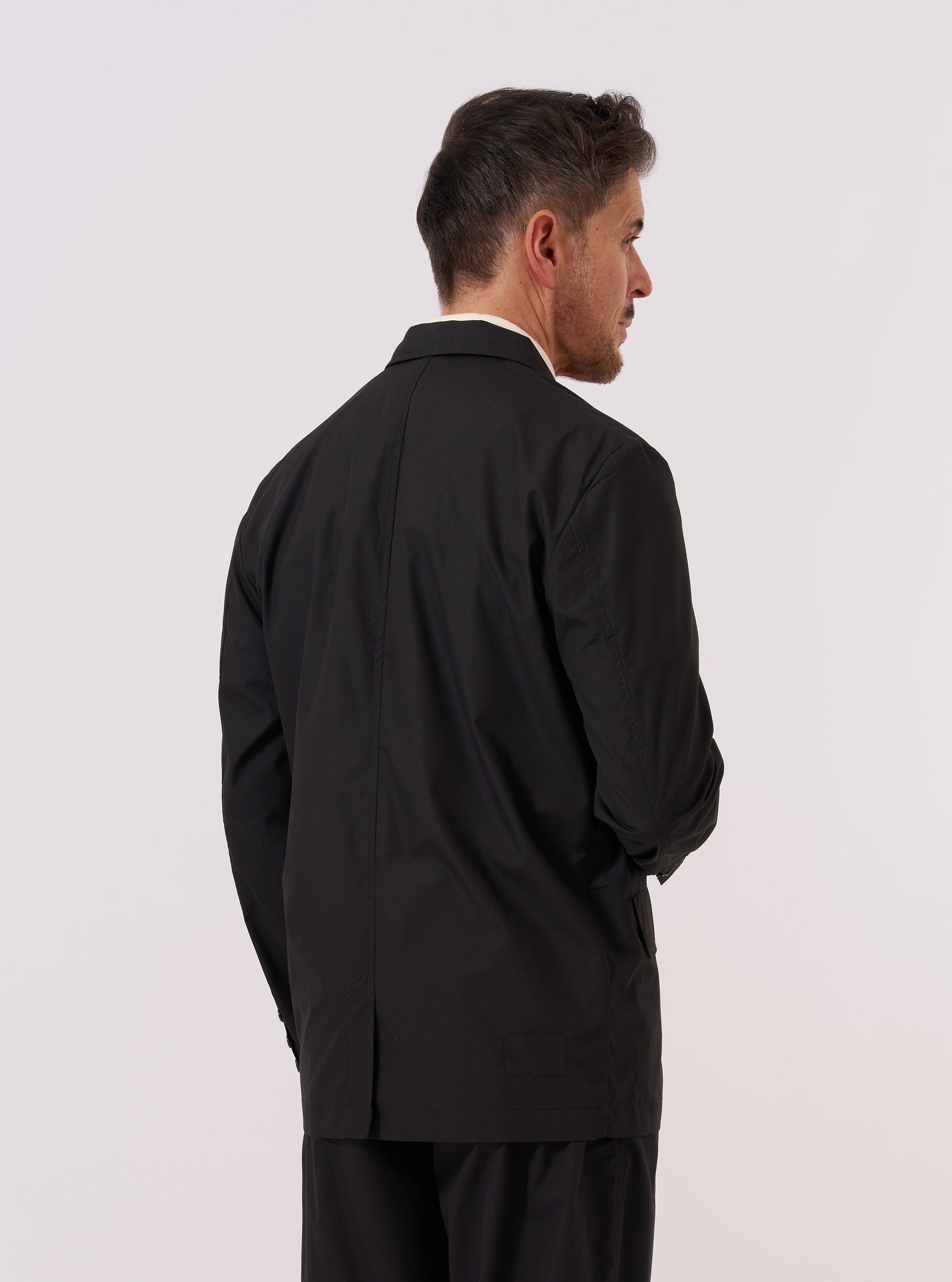 A model wears the Black Recycled Polytech Capitol Jacket over a light-colored top, shown from a rear three-quarter angle against a plain, neutral studio background. The jacket features a single back vent, structured seams, and fully functioning cuffs. The black recycled polytech fabric has a smooth, technical finish.