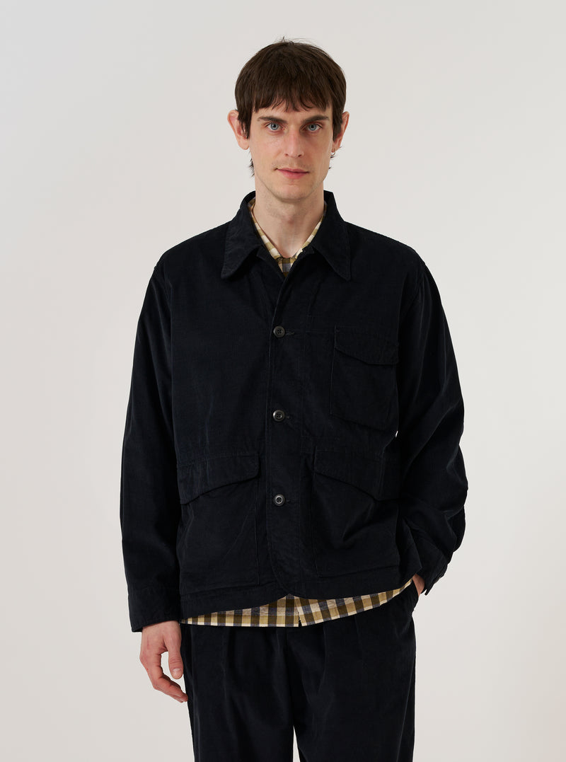 A model wears the Black Organic Fine Cord Duty Jacket over a white t-shirt, standing against a plain white background. The jacket features four front patch pockets, a structured collar, and a central button closure. The black organic cotton corduroy fabric has a visible fine-ribbed texture.