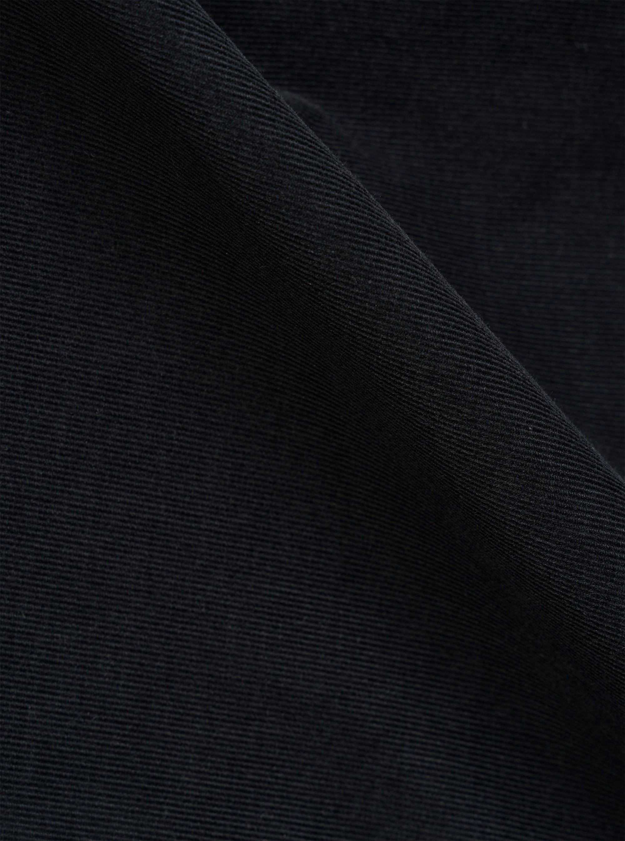 A close-up swatch image of the Black Organic Fine Cord Duty Jacket fabric highlights the fine-wale texture of the black corduroy material. The vertical ridges of the organic cotton fabric are clearly visible, filling the entire frame to show the garment's detailed construction and color.