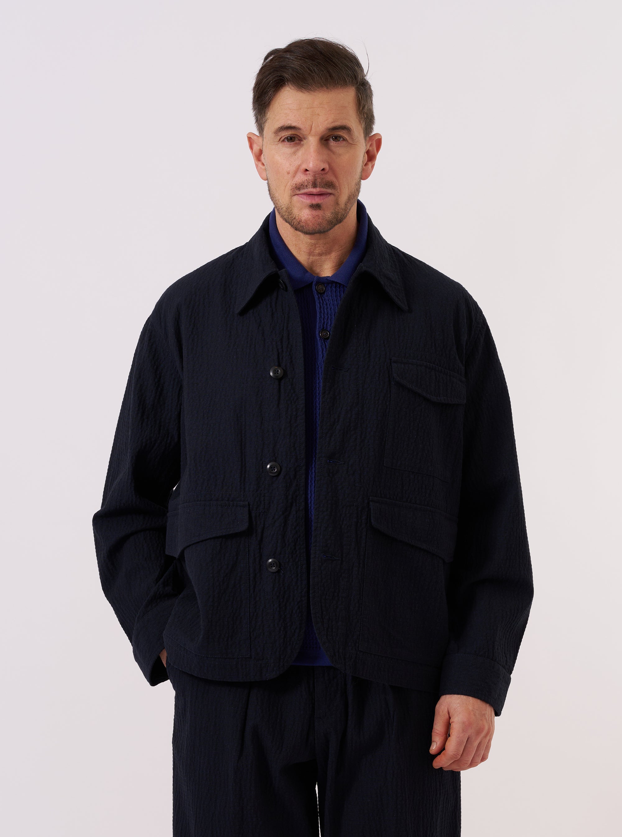 A model wears the Dark Navy Ospina Cotton Duty Jacket over a light-colored top, standing against a plain, neutral background. The jacket features a button-front closure, a structured collar, and multiple large patch pockets on the front. The fabric displays a subtle, puckered seersucker texture characteristic of the Ospina cotton.