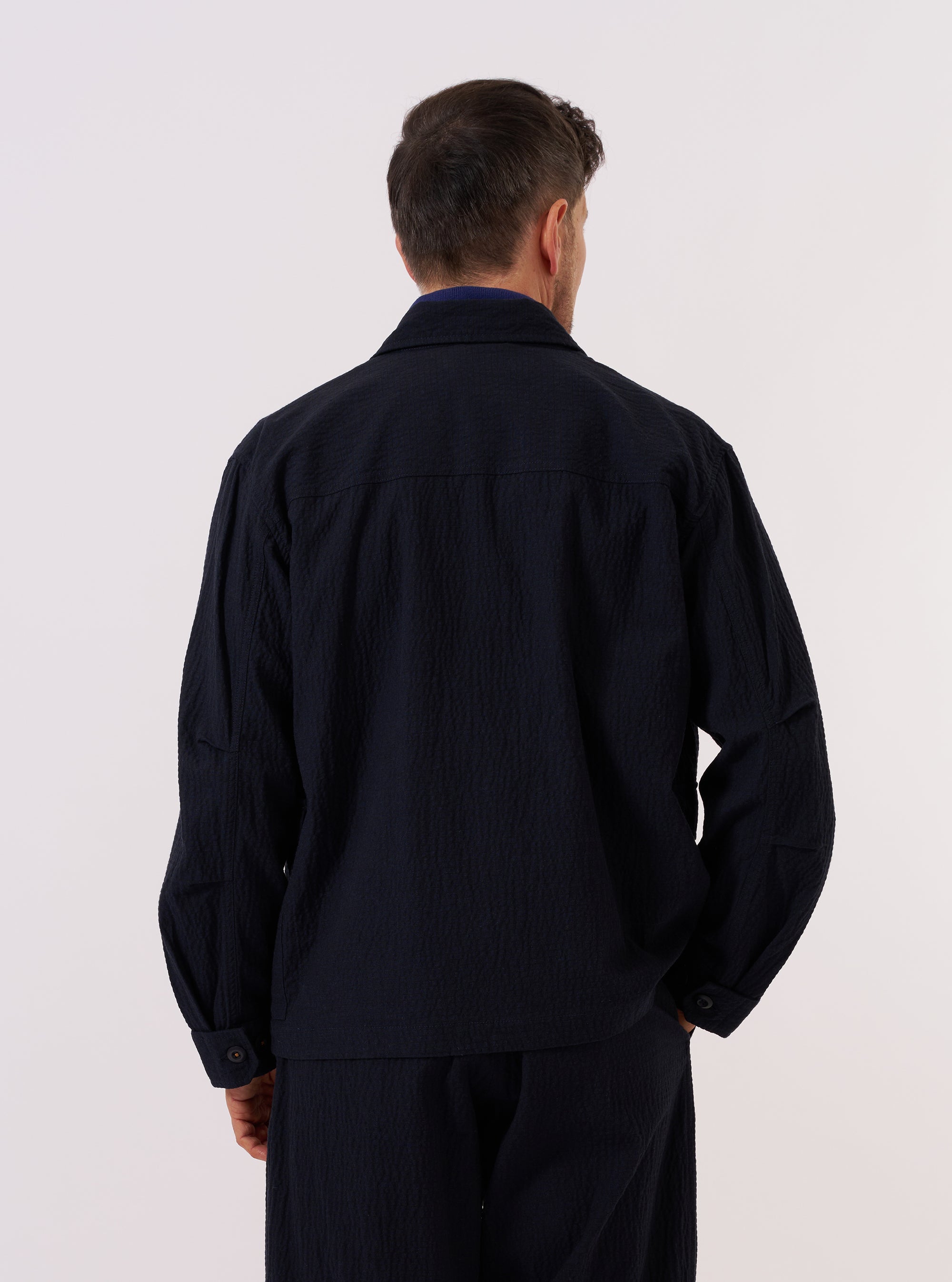 A model wears the Dark Navy Ospina Cotton Duty Jacket over a light-colored shirt, shown from a rear three-quarter angle against a plain, neutral studio background. The jacket features a structured collar and a straight hem, highlighting the distinct puckered texture of the dark navy seersucker fabric.